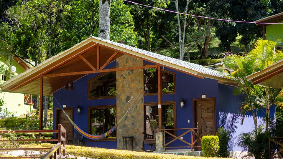 Cabaña de Villa Shalom en Santa Rosa de Copán, Honduras, con fachada azul, detalles en piedra y hamaca en el exterior rodeada de árboles.