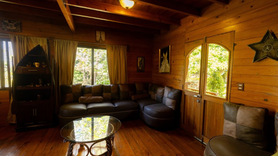 Sala acogedora en una cabaña de Villa Shalom, con sofá negro, mesa de vidrio y ventanas que permiten la entrada de luz natural.