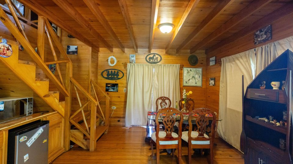 Interior de una cabaña en Villa Shalom, con paredes de madera, una mesa con sillas y escaleras de acceso al segundo nivel.