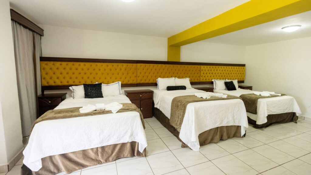 Habitación en Villa Shalom con tres camas matrimoniales, decoradas con sábanas blancas y toallas, y un ambiente acogedor.