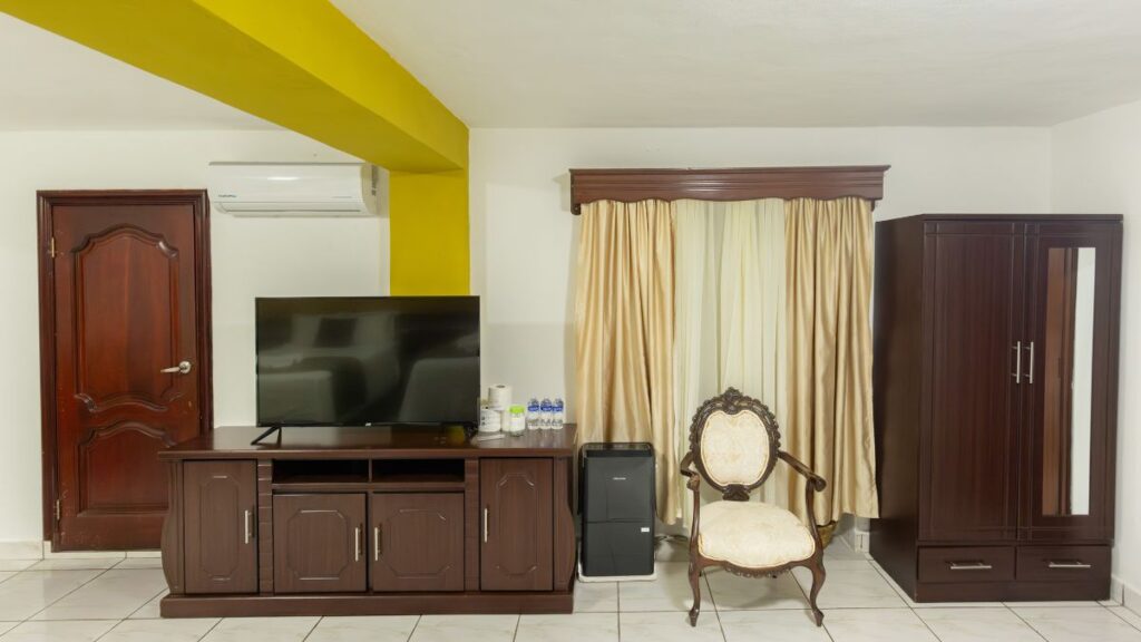 Habitación en Villa Shalom con un mueble de madera, televisor, silla elegante y cortinas beige. Ambiente acogedor y luminoso.