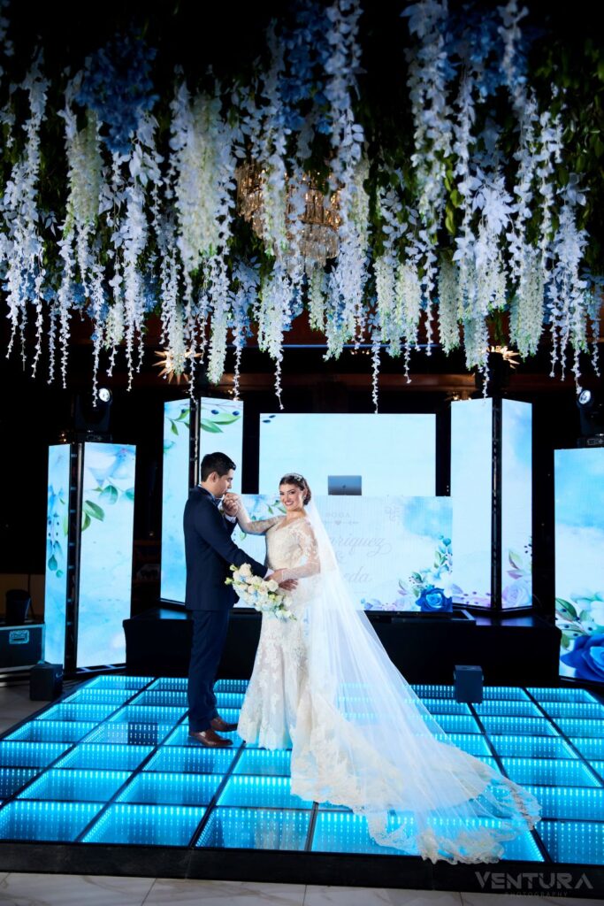 Pareja de novios intercambiando votos en una ceremonia de boda en Villa Shalom, decorada con flores colgantes y pantallas iluminadas.