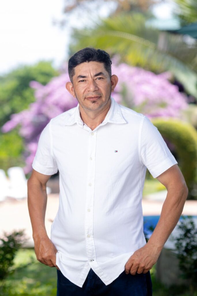 Hombre de pie con camisa blanca, sonriendo frente a un fondo de flores moradas en Villa Shalom, Santa Rosa de Copán, Honduras.