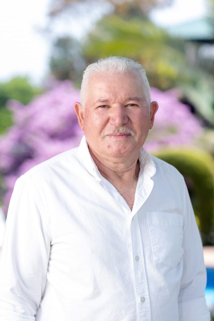 Retrato de un hombre mayor con camisa blanca, sonriendo frente a un fondo de flores moradas en Villa Shalom, Santa Rosa de Copán, Honduras.