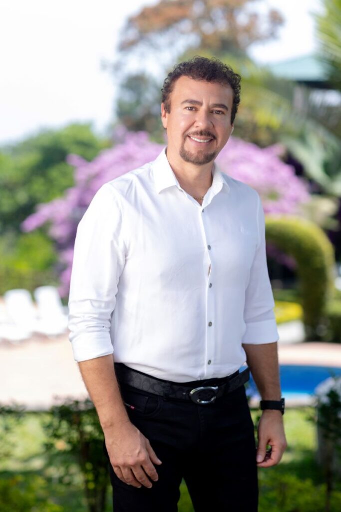 Hombre sonriendo con camisa blanca y pantalón oscuro, posando en los jardines de Villa Shalom en Santa Rosa de Copán, Honduras.