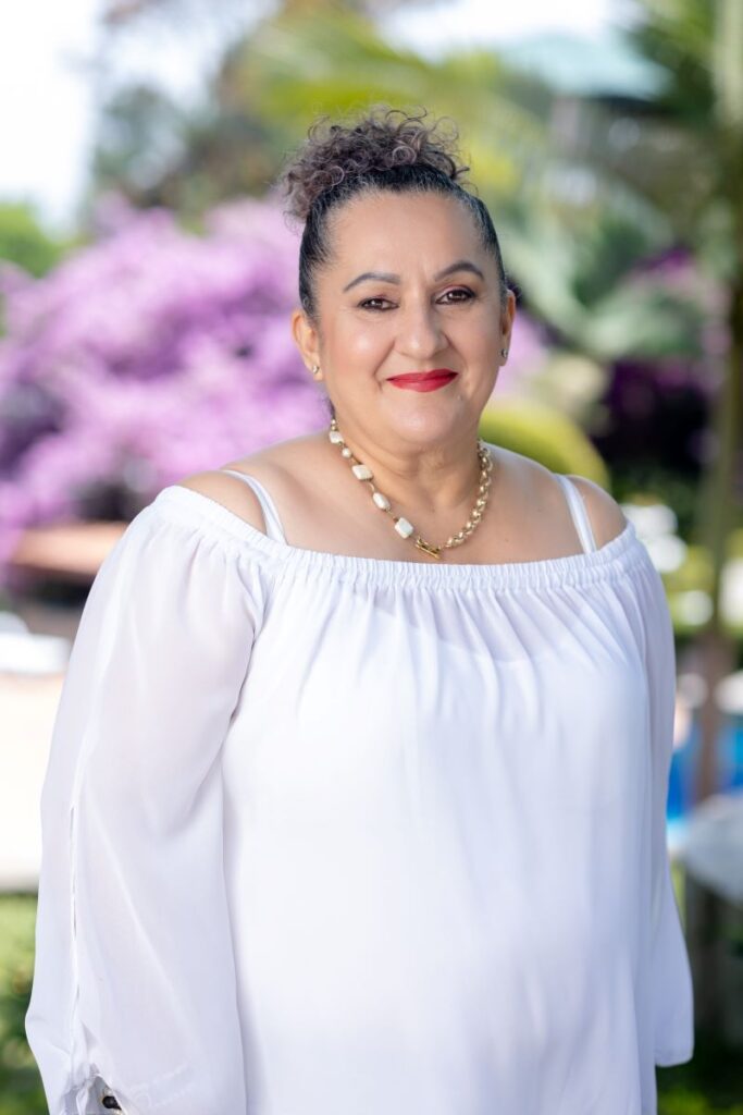 Mujer sonriente vestida de blanco, con joyas, en un entorno natural con flores moradas, en Villa Shalom, Santa Rosa de Copán, Honduras.